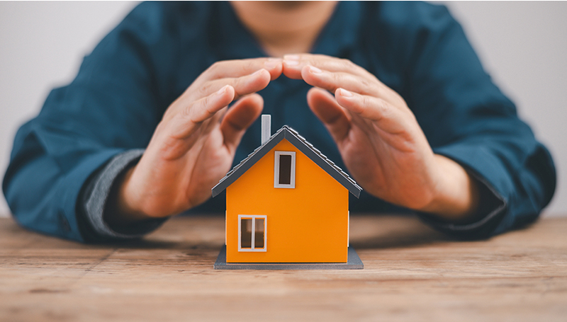 Hands forming a protective roof shape over a miniature orange house